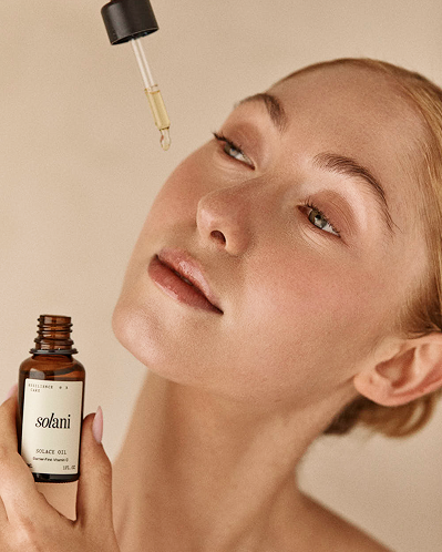 Model holding Solani Solace Oil with the glass dropper raised, a drop of oil falling